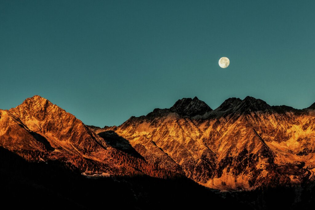 A stunning view of rugged mountain peaks illuminated by the warm glow of sunset, with a full moon rising in the clear evening sky.